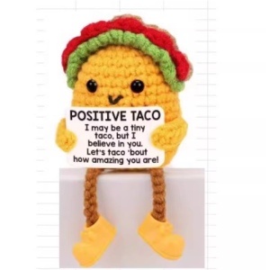 Hand-crocheted Taco doll holding a white positive affirmation sign, sitting on a wooden table.