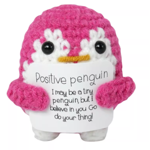 Hand-crocheted penguin doll holding a white positive affirmation sign, sitting on a wooden table.