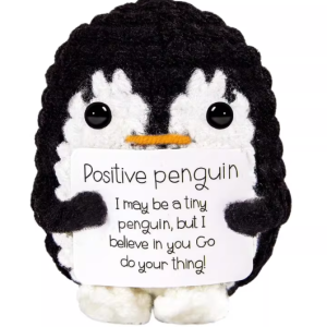 Hand-crocheted penguin doll holding a white positive affirmation sign, sitting on a wooden table.