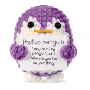 Hand-crocheted penguin doll holding a white positive affirmation sign, sitting on a wooden table.
